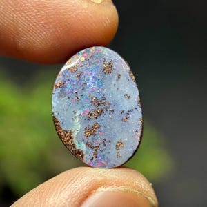 May include: Oval-shaped opal gemstone with a light blue base color and iridescent flashes of blue, red, and green. The opal has brown matrix inclusions and is held between two fingers.