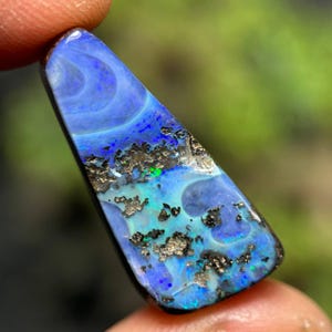 May include: A polished, elongated opal gemstone with a vibrant display of blues, greens, and black matrix. The stone's surface showcases swirling patterns and flashes of color, creating a captivating visual effect. The opal is held by a finger.