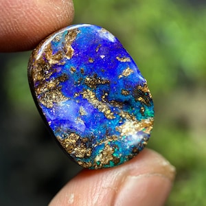 May include: A vibrant, polished opal gemstone with a striking blue base color, accented by shimmering gold and hints of green. The oval-shaped stone is held between fingers, showcasing its natural beauty and intricate patterns. A unique piece for jewelry or collection.