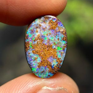 May include: Oval-shaped opal gemstone with a vibrant display of colors. The stone features a mix of blue, green, and gold hues, with a speckled pattern. The opal is held between two fingers, showcasing its natural beauty and unique characteristics.