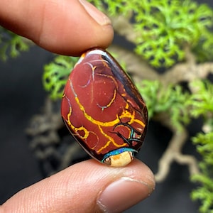 May include: A polished, oval-shaped gemstone with a deep red base color, featuring intricate patterns of gold, black, and blue. The gemstone is held between fingers, showcasing its unique color variations and textures. The stone is likely an opal.