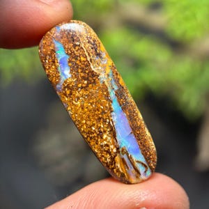 Könnte beinhalten: Ein länglicher, polierter Edelstein mit einer erdigen Farbpalette. Der Opal zeigt eine gesprenkelte, goldbraune Basis mit lebendigen Streifen in Blau und Türkis. Der Stein wird zwischen den Fingern gehalten.