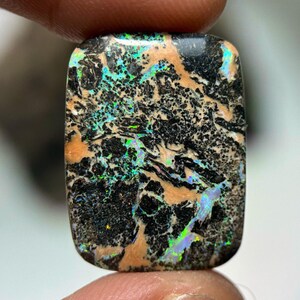 Puede incluir: Una gema rectangular con una superficie pulida. La piedra presenta un patrón complejo de colores negro, beige y verde y azul iridiscente. La gema se sostiene entre dos dedos.