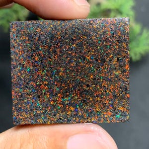 May include: A square-shaped piece of dark, textured material featuring a dense distribution of iridescent flecks in shades of orange, red, green, and blue. The surface has a granular, matte appearance with a consistent pattern of colourful light reflections.