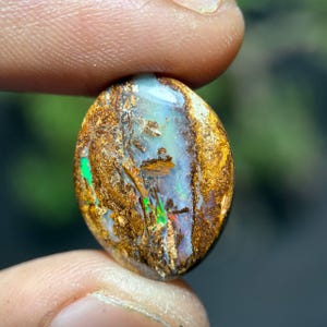 May include: Oval-shaped opal gemstone with a mix of colors. The opal has a brown matrix with flashes of green, blue, and red. The gemstone is held between two fingers, showcasing its natural beauty and unique patterns.