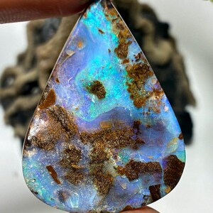 May include: A teardrop-shaped opal gemstone with a vibrant display of colors. The stone features a base of light blue and purple hues, accented with patches of brown and areas of iridescent green. The opal is held up by a hand.