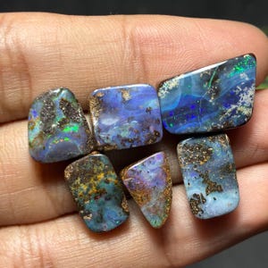 May include: A collection of six polished opal gemstones. The opals display a variety of colors, including blues, greens, and purples, with iridescent flashes. Each stone has a unique pattern and shape, with a dark matrix.