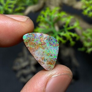 May include: A colorful opal gemstone with a triangular shape. The stone displays a mix of colors, including green, blue, and gold, with a white base and brown accents. The opal is held between two fingers.