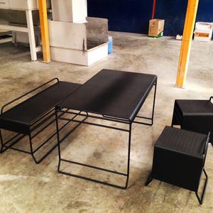 Puede incluir: Juego de muebles de exterior de metal negro. Incluye una mesa rectangular, un banco y dos taburetes cuadrados. La mesa y el banco tienen una superficie perforada. El conjunto tiene un diseño minimalista.