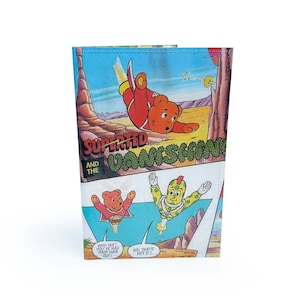 May include: A wallet featuring a comic book illustration of a cartoon bear in a red suit, with the text "SUPERTED AND THE VANISHING". The comic strip style design includes speech bubbles and a desert landscape background.