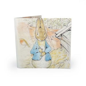 May include: A tri-fold wallet featuring a watercolor illustration of Peter Rabbit in a blue jacket. The background includes a garden scene with pots and a bird. The wallet is white with a printed design.