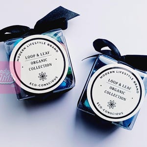 May include: Two clear, square boxes filled with colourful sweets, each tied with a black ribbon. The boxes have a circular label that reads "LOOP & LEAF ORGANIC COLLECTION" and "ECO-CONSCIOUS". The boxes are on a white surface.
