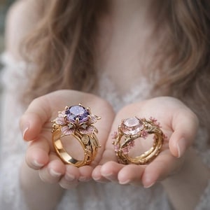 Op de afbeelding: Twee sierlijke gouden ringen. Eén ring heeft een grote paarse edelsteen in een bloemdesign met roze emaille accenten. De andere ring heeft een roze edelsteen en is omgeven door delicate roze bloemen.