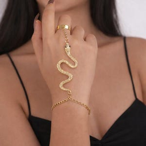 May include: Gold-coloured snake hand chain jewellery. The ring connects to a snake-shaped chain that drapes across the hand and connects to a bracelet. The snake has black eyes and a textured body. The jewellery is worn on a hand with a black top in the background.