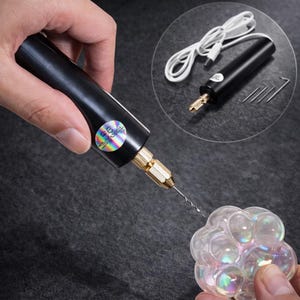 Op de afbeelding: Een zwarte mini-boormachine met een goudkleurige punt wordt in een hand gehouden en boort in een helder, iriserend object. Een close-up toont de boormachine, een witte USB-kabel en kleine gereedschappen. De boormachine heeft een holografische sticker.