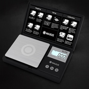 May include: A black digital pocket scale with a stainless steel weighing surface. The scale has a digital display showing "0.00" and the brand name "MAXUS". Instructions for weighing, taring, and calibration are printed on the lid.