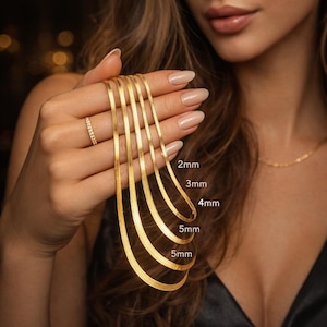 May include: Several gold herringbone chain necklaces, ranging from 2mm to 5mm in width, are displayed. The image shows the different chain sizes, with text indicating the measurements. The necklaces have a reflective surface and are set against a dark backdrop.