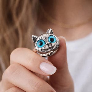 May include: A silver cat ring with large, bright blue eyes. The ring features a detailed cat face with pointed ears and a smiling mouth. The ring is held up by a hand.