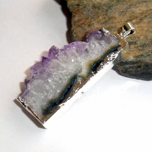 Puede incluir: Un colgante de plata con un cristal de amatista en bruto con un degradado de color p&uacute;rpura y blanco. El cristal tiene un borde natural y dentado y est&aacute; engastado en un marco de plata.