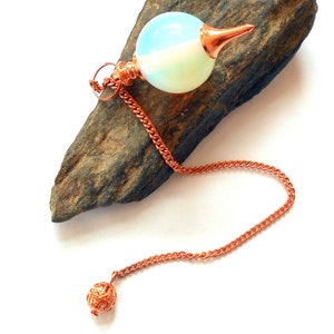 May include: A white and blue opal pendulum with a copper chain and a copper ball at the end. The pendulum is resting on a piece of brown rock.