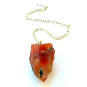 May include: A carnelian crystal pendulum with a silver chain. The crystal is a deep orange color and has a rough, natural shape.
