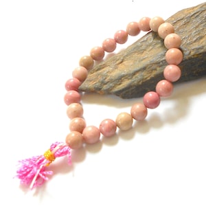 Puede incluir: Un brazalete de piedras preciosas rosa y blanco con un borla rosa. El brazalete está hecho de cuentas redondas y está enhebrado en un cordón delgado.