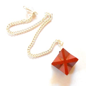 Puede incluir: Un péndulo de cristal rojo con una cadena de plata. El cristal tiene forma de pirámide con una base cuadrada.