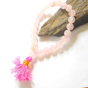 Puede incluir: Un brazalete de cuentas de cuarzo rosa con un borla rosa. El brazalete está hecho de cuentas redondas y está enhebrado en un cordón elástico transparente.