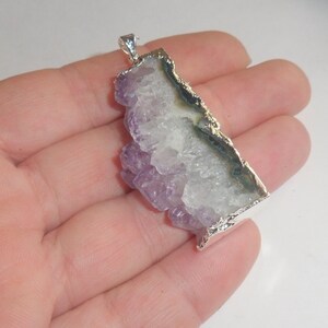 Puede incluir: Un colgante de plata con un gran cristal de amatista con un borde natural y sin pulir. El cristal es de color p&uacute;rpura claro y tiene un centro blanco claro. El colgante est&aacute; engastado en un marco de plata con un borde texturizado.