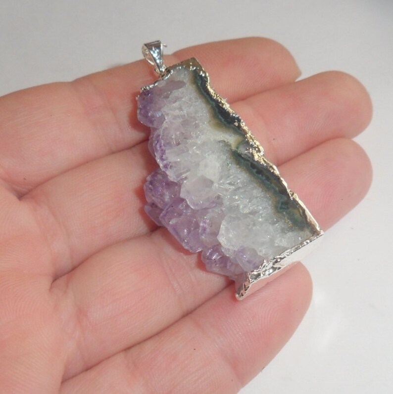 Puede incluir: Un colgante de plata con un gran cristal de amatista con un borde natural y sin pulir. El cristal es de color p&uacute;rpura claro y tiene un centro blanco claro. El colgante est&aacute; engastado en un marco de plata con un borde texturizado.