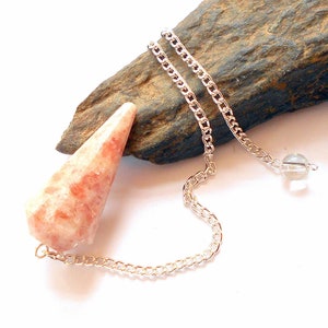 May include: A sunstone pendulum with a silver chain and a clear crystal bead at the end. The pendulum is a teardrop shape and has a light orange and white color.