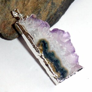 Puede incluir: Un colgante de plata con un gran cristal de amatista con una inclusi&oacute;n verde y dorada. El cristal tiene una textura rugosa y natural y est&aacute; engastado en un marco de plata.