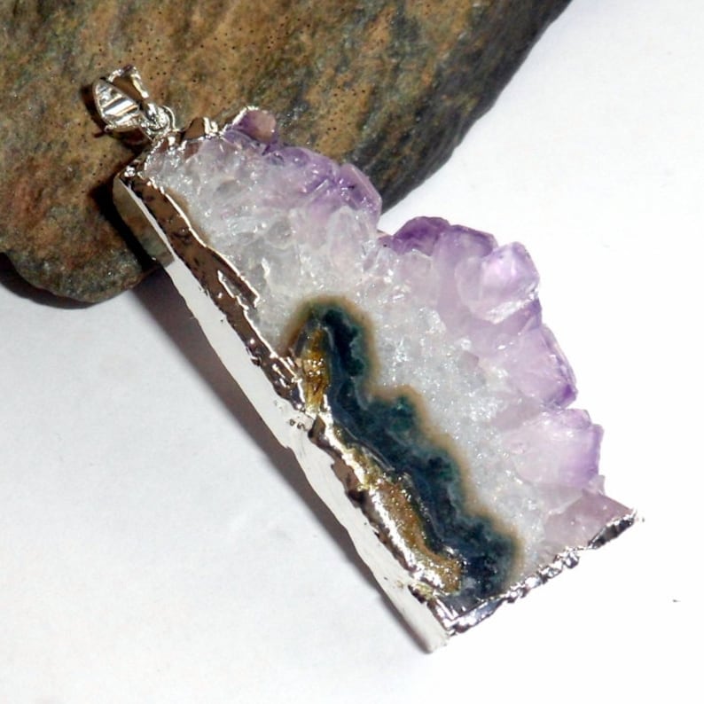 Puede incluir: Un colgante de plata con un gran cristal de amatista con una inclusi&oacute;n verde y dorada. El cristal tiene una textura rugosa y natural y est&aacute; engastado en un marco de plata.