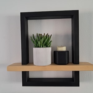 May include: A contemporary wall-mounted shelf unit with a black frame and a natural wood shelf. It displays a white and grey striped planter with a green succulent, and a black geometric candle holder with a cream candle.