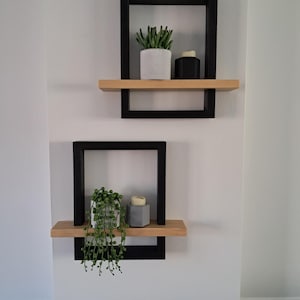 May include: Two contemporary floating shelves fixed to a white wall. Each shelf features a black frame, a natural wood shelf, and is adorned with a small potted plant and a decorative item. The shelves are positioned at intervals.