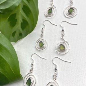May include: Three pairs of silver-toned drop earrings, each featuring a small, irregular piece of translucent olive green sea glass set within a spiralling wire frame. The earrings hang from standard silver-toned ear wires with a small link chain.
