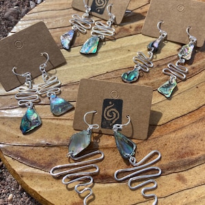 May include: Three pairs of silver-toned wire earrings displayed on brown cardboard tags. Each earring features a zigzag or coiled wire design paired with an iridescent, multi-coloured abalone shell fragment. The earrings hang from silver-toned hooks.