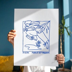 May include: A white print with a blue line drawing of a person with an umbrella, a flower, and a puddle. The text "3:01 PM Puddle On Through" is at the bottom. The print is held by a person.