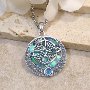 May include: A circular silver pendant on a chain featuring an iridescent abalone shell inlay. The center displays an intricate Celtic knot design, while the outer border is engraved with small crescent moons and stars, accented by a single blue stone.