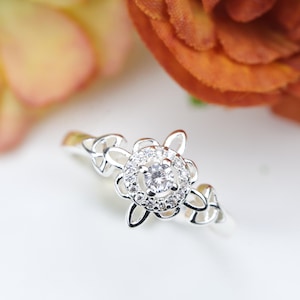 Peut inclure: Une bague en argent avec une pierre centrale ronde et transparente entourée d'un halo de pierres plus petites. Le sertissage en forme de fleur présente des boucles métalliques ajourées sur l'anneau, créant un design détaillé et élégant.