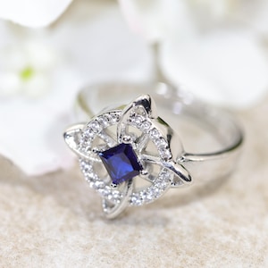 May include: A silver-toned ring featuring a central square-cut deep blue gemstone surrounded by a circular frame of small clear pavé stones. The metal setting forms a compass-like design with four pointed extensions radiating from the centre.