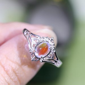 May include: A silver ring with an oval-shaped, multi-colored gemstone. The gemstone has shades of orange, yellow, and clear. The ring has an ornate, filigree design around the gemstone and on the band. The ring is held by a person.