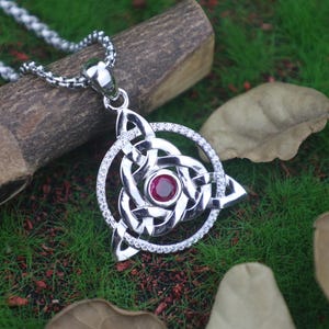 Könnte beinhalten: Silberfarbene Keltischer Knoten-Anhänger-Halskette mit einem roten Edelstein in der Mitte. Der Anhänger hat ein detailliertes, verschlungenes Design, umgeben von einem Ring aus kleinen, klaren Steinen. Die Halskette ist silberfarben.