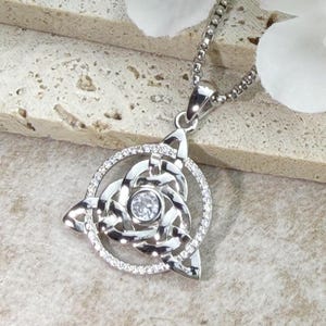 Pode incluir: Um colar com pendente em tom prateado com uma moldura circular incrustada com pequenas pedras claras. O centro contém um intrincado design de nó celta triquetra em metal polido com uma pedra redonda clara no meio.