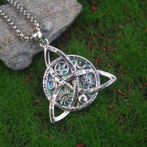 May include: Silver-toned pendant necklace with a Celtic knot design. The pendant features an abalone inlay and engraved text including "KNOW YOUR POWER" and "BLESSED BE". The necklace rests on a bed of green grass.