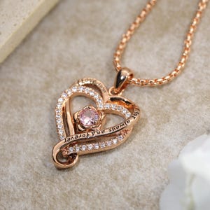 May include: A rose gold-toned heart pendant necklace featuring a central round pink stone and a border of small clear crystals. The metal frame is engraved with the text "for always and forever" and hangs from a matching rose gold-toned chain.