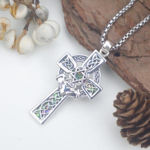 Puede incluir: Colgante de cruz celta plateada con intrincados nudos e incrustaciones de concha de abulón iridiscente. El colgante cuelga de una cadena plateada. El diseño de la cruz presenta un círculo central y cuatro brazos con patrones detallados.