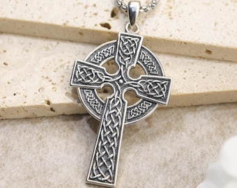 May include: A silver-toned metal Celtic cross pendant featuring intricate, embossed knotwork patterns throughout the cross and the surrounding circle. The pendant is suspended from a matching silver-toned metal chain with a textured, rounded link design.