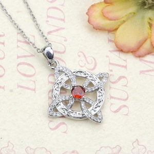 May include: A silver-toned pendant necklace featuring a Celtic knot design. The pendant has a central red gemstone and is surrounded by clear, sparkling stones. The necklace is displayed on a patterned background with a flower in the upper right corner.