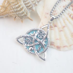 May include: A silver-toned metal pendant necklace featuring a triquetra shape surrounding a circular light blue inlay. The metal surface is engraved with symbols including a pentagram, crescent moons, and an eye, along with text: SPEAK WITH LOVE, BE KIND, BLESSED BE, BE BALANCED, HONOR YOUR WORD, KNOW YOUR POWER, LISTEN WITH LOVE, MIND BOUNDARIES.